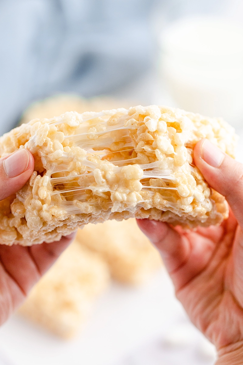Hand holding a half batch homemade Rice Krispie treat with visible stretchy marshmallow strands.