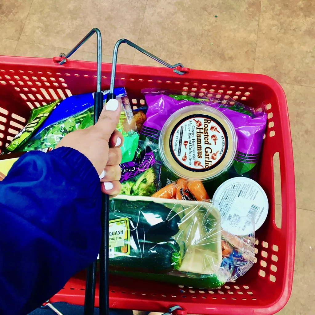A person pushing a red shopping basket filled with various vegetables and packaged food items.